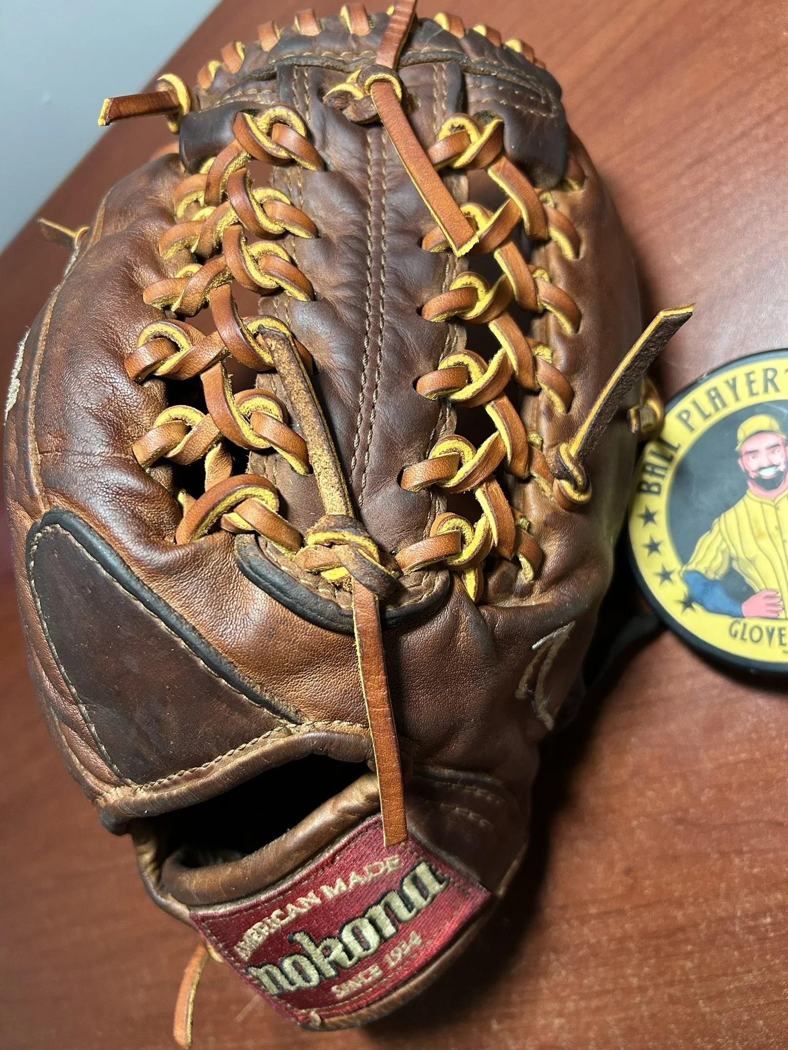 Old brown leather baseball glove with yellow laces placed on a wooden surface, with a round patch that reads "Ball Player Glove" featuring a cartoon character wearing a baseball cap and uniform.