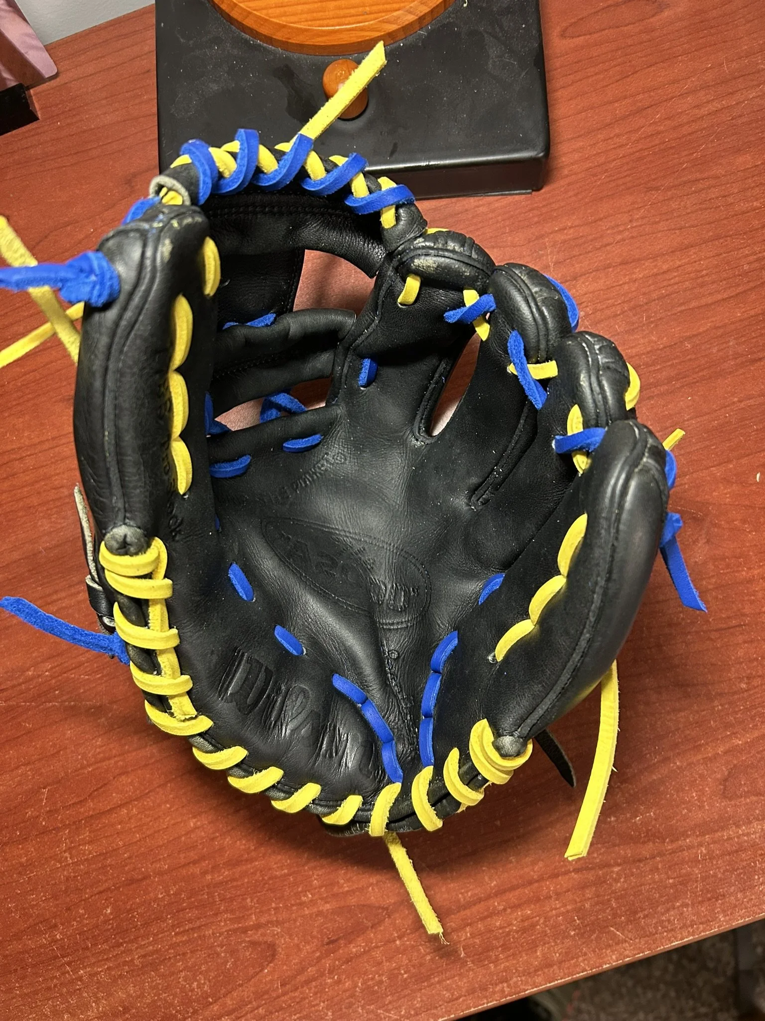 A black baseball glove with yellow and blue laces on a wooden surface, with a scale in the background.