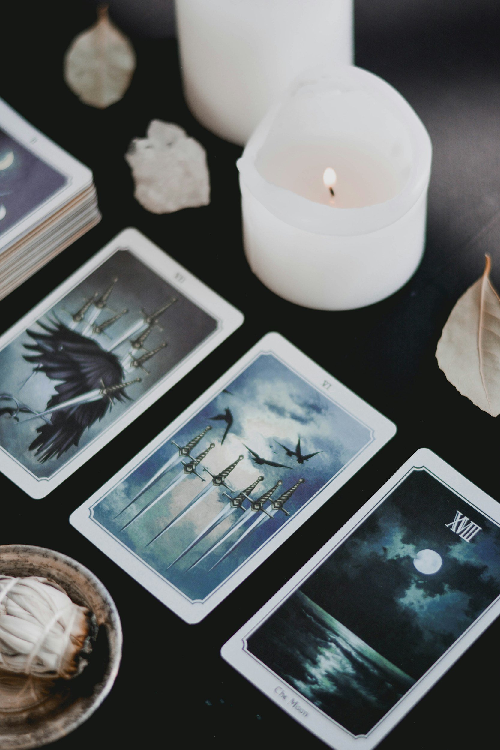 Tarot cards laid out on a black surface with a lit white candle and some shells or stones around, along with a dried leaf and a small dish.