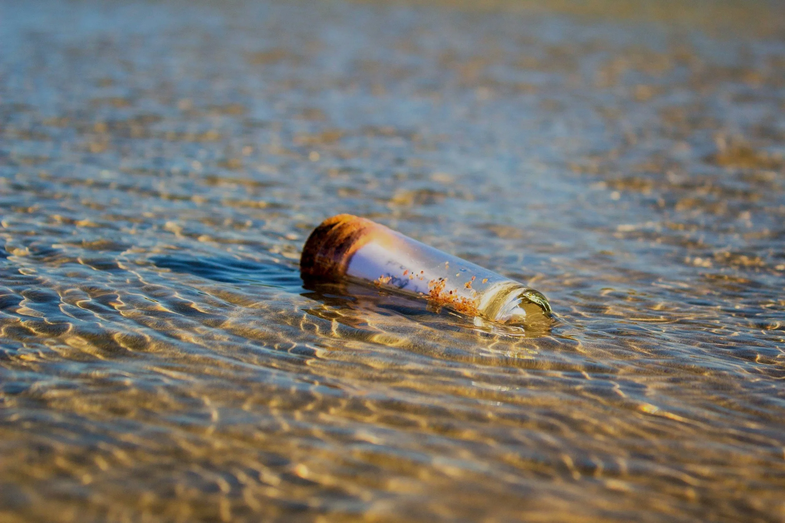 A glass bottle with a cork, partly submerged in shallow water with ripples, on a sandy beach.
