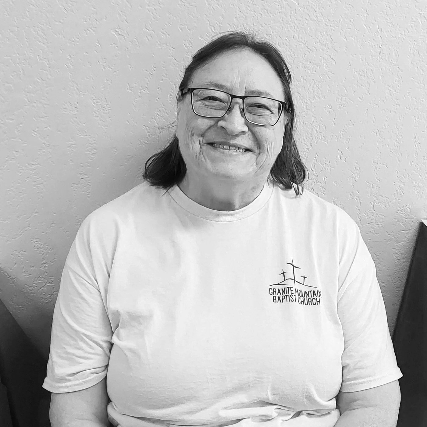 A smiling woman with glasses and shoulder-length hair wearing a white T-shirt with a logo that says 'Granite Mountain Baptist Church,' sitting against a textured wall.