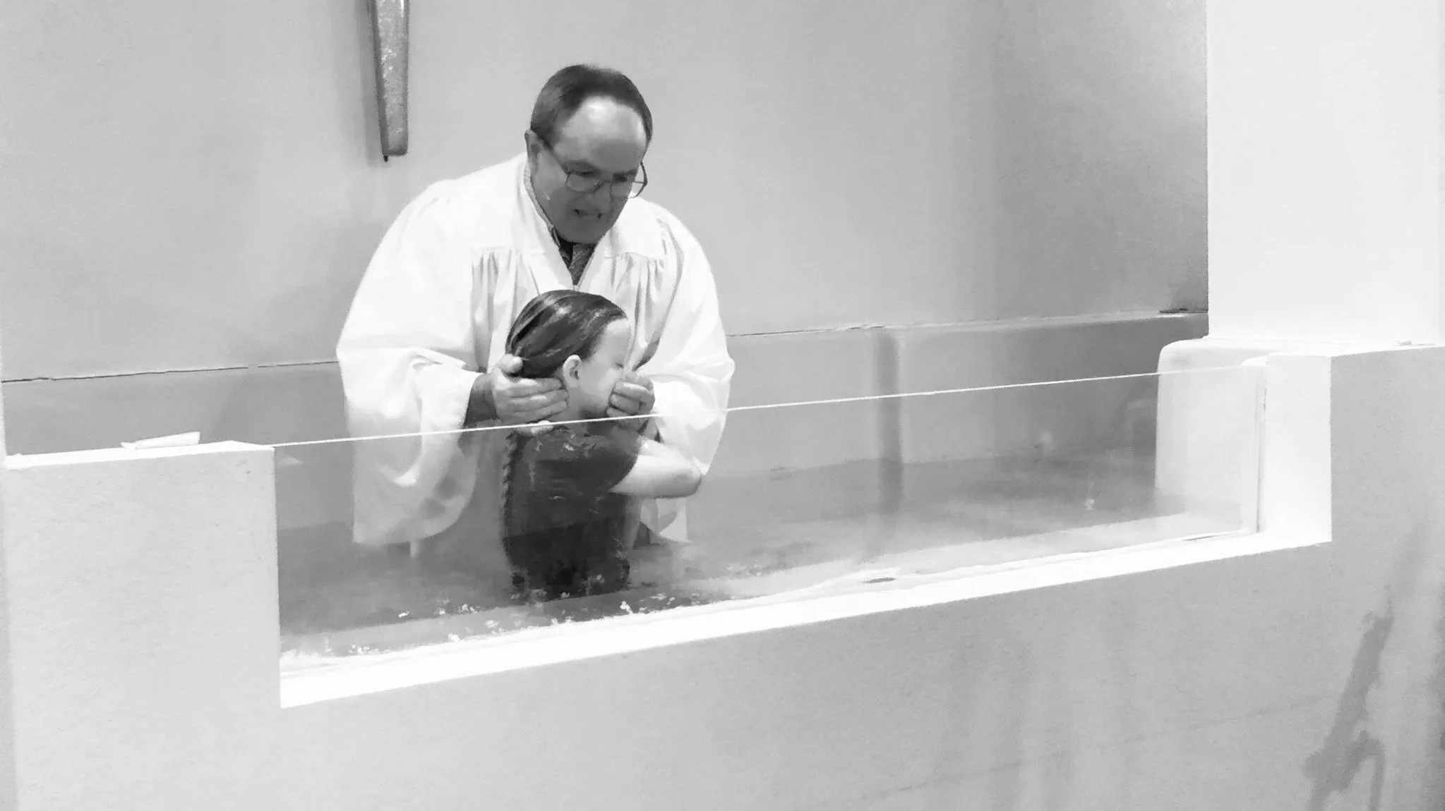 A man in a white robe baptizing a woman submerged in water, holding her head and neck during the baptism.