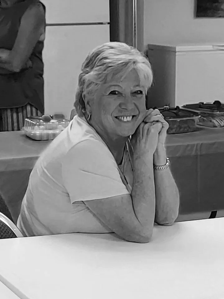 A smiling elderly woman with short hair sitting at a table, resting her chin on her hands with a watch on her wrist, in an indoor setting.