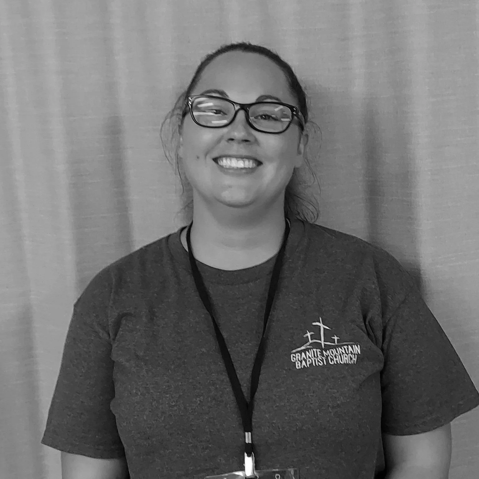 A smiling woman wearing glasses and a Granite Mountain Baptist Church T-shirt, standing in front of a curtain.