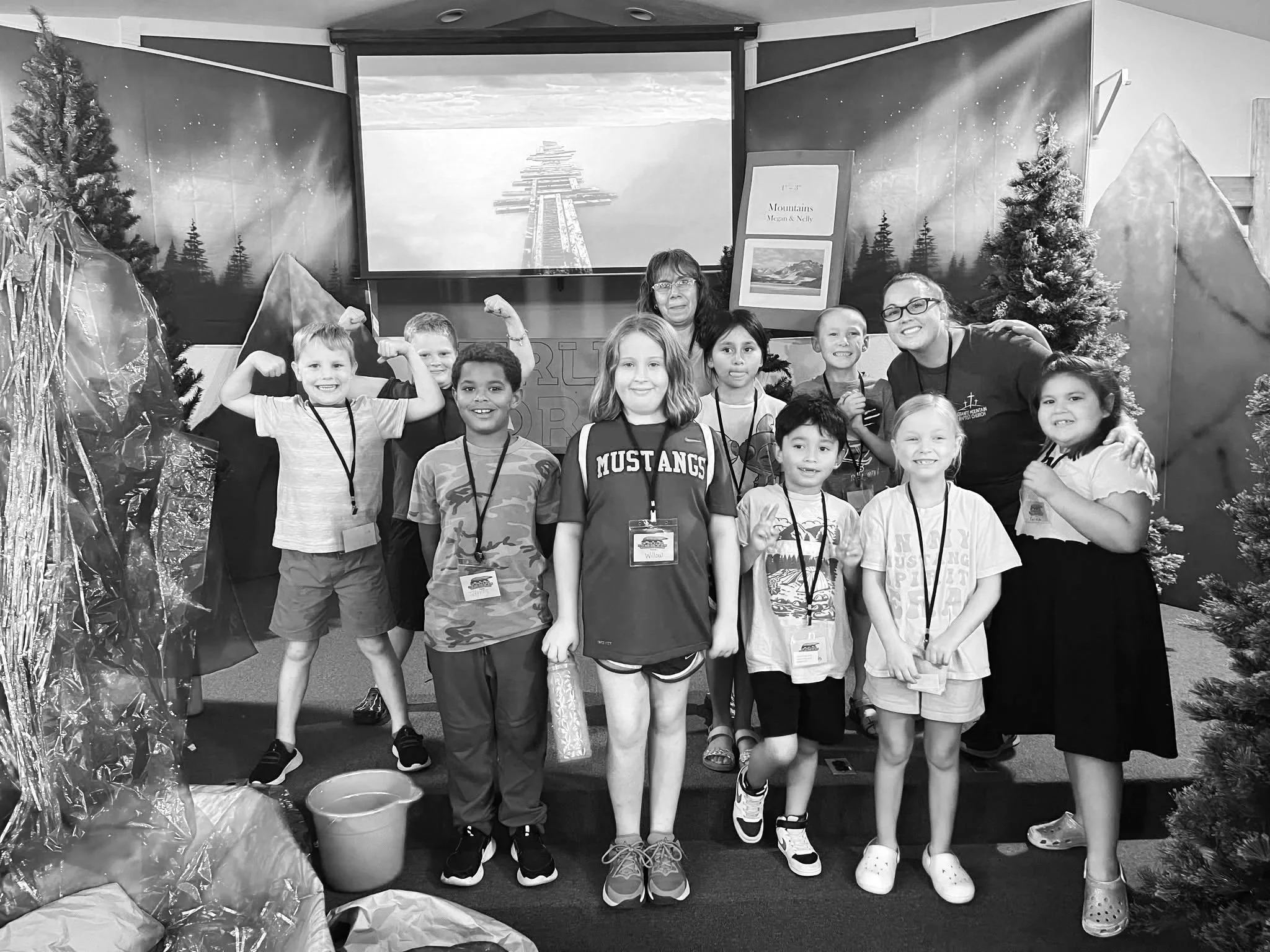 Group of children and two adults standing in a room with a backdrop of mountains, trees, and a lighthouse. The children are smiling, some posing with their arms flexed. The scene appears to be a school or camp activity.