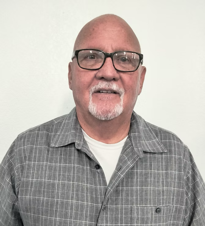 A bald man with glasses, a white goatee, wearing a gray plaid shirt over a white shirt, standing against a plain white background.