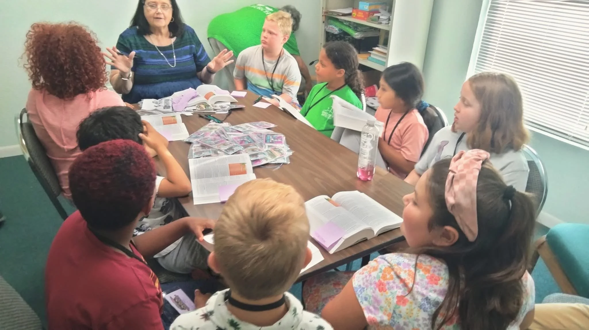 A group of children and an adult woman seated around a table engaged in a discussion or learning activity, with books and papers spread across the table.