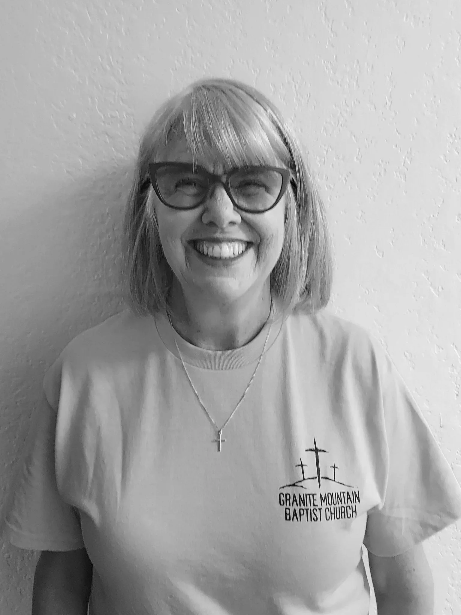 A smiling woman with glasses and shoulder-length hair wearing a T-shirt with 'Granite Mountain Baptist Church' logo, standing against a textured wall.
