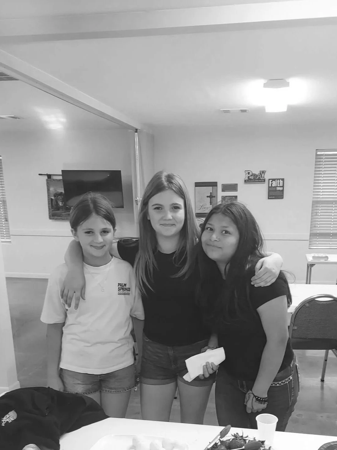 Three young girls standing close together with their arms around each other, smiling in a room with wall decorations and a TV in the background.