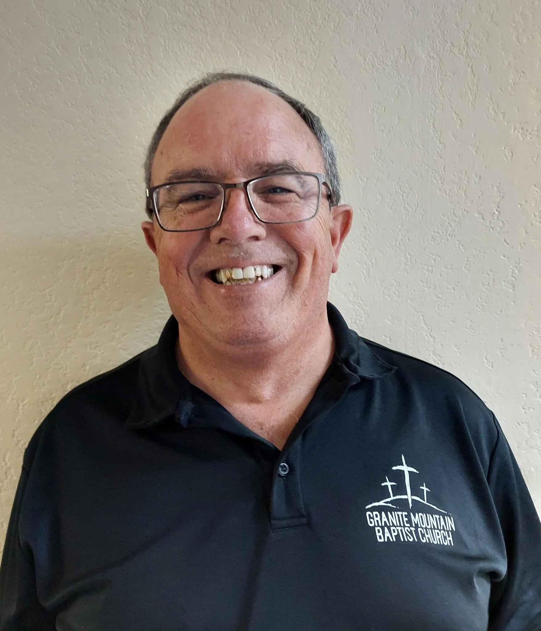 A smiling man with glasses and a black polo shirt with the logo of Granite Mountain Baptist Church, standing against a beige textured wall.