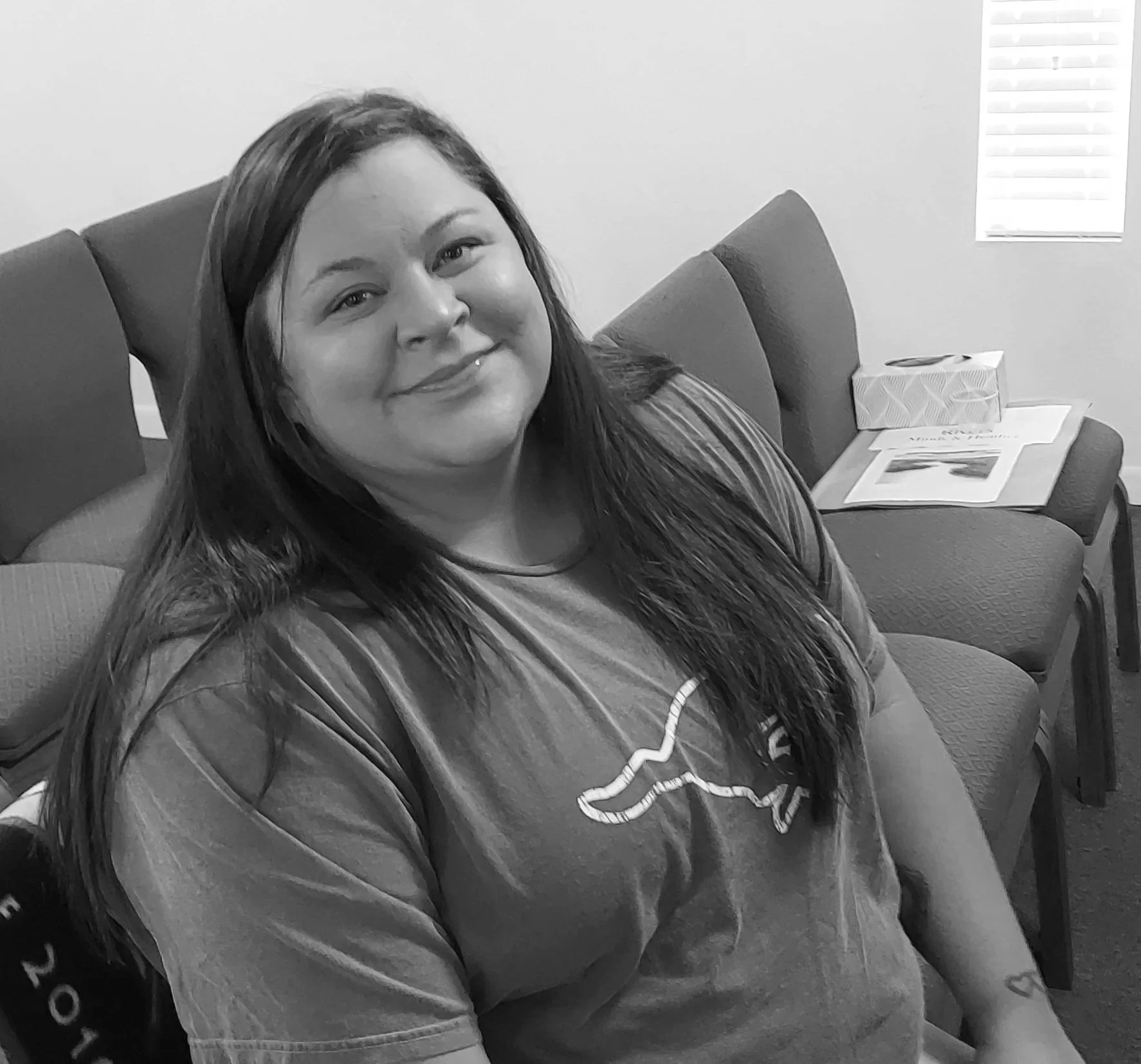 A smiling woman with long hair sitting on a chair in a room, with a tissue box and papers on a table behind her.