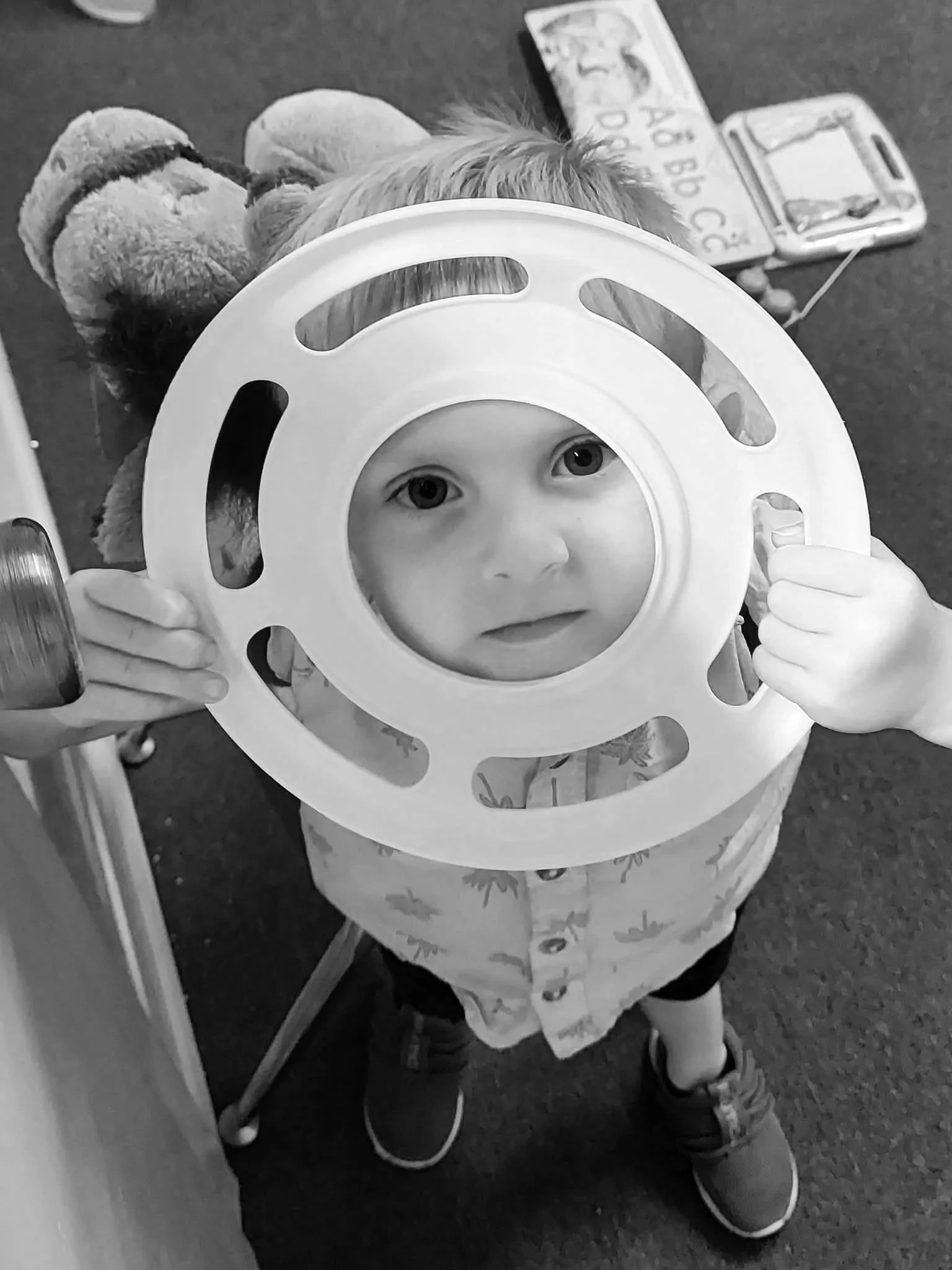 A young child looking through a circular toy ring, holding it in front of their face, in a room with toys and educational materials on the floor.