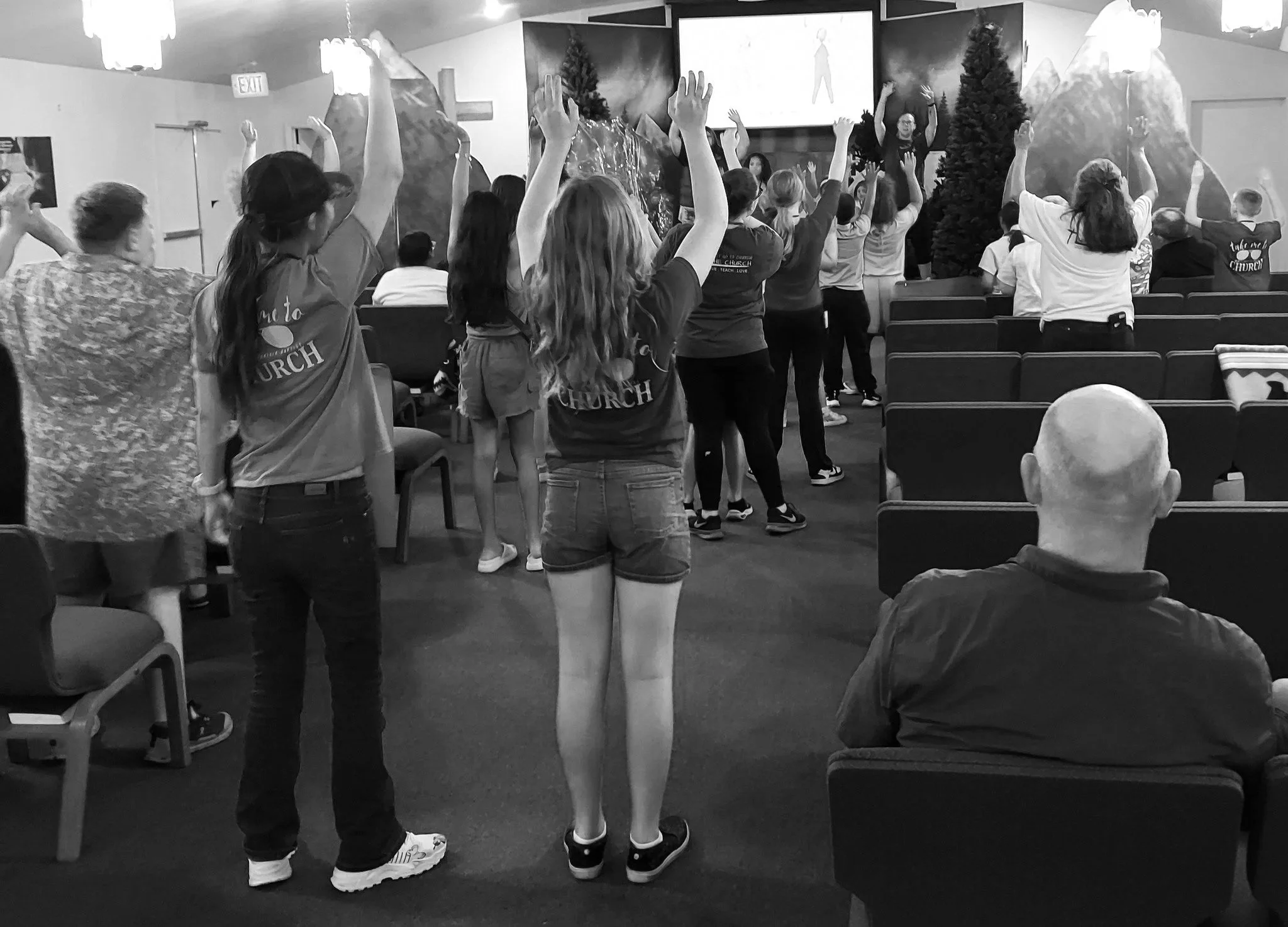 People in a church engaging in a group activity with their hands raised. The church features a cross on the wall, some Christmas trees, and mountain scenery artwork.