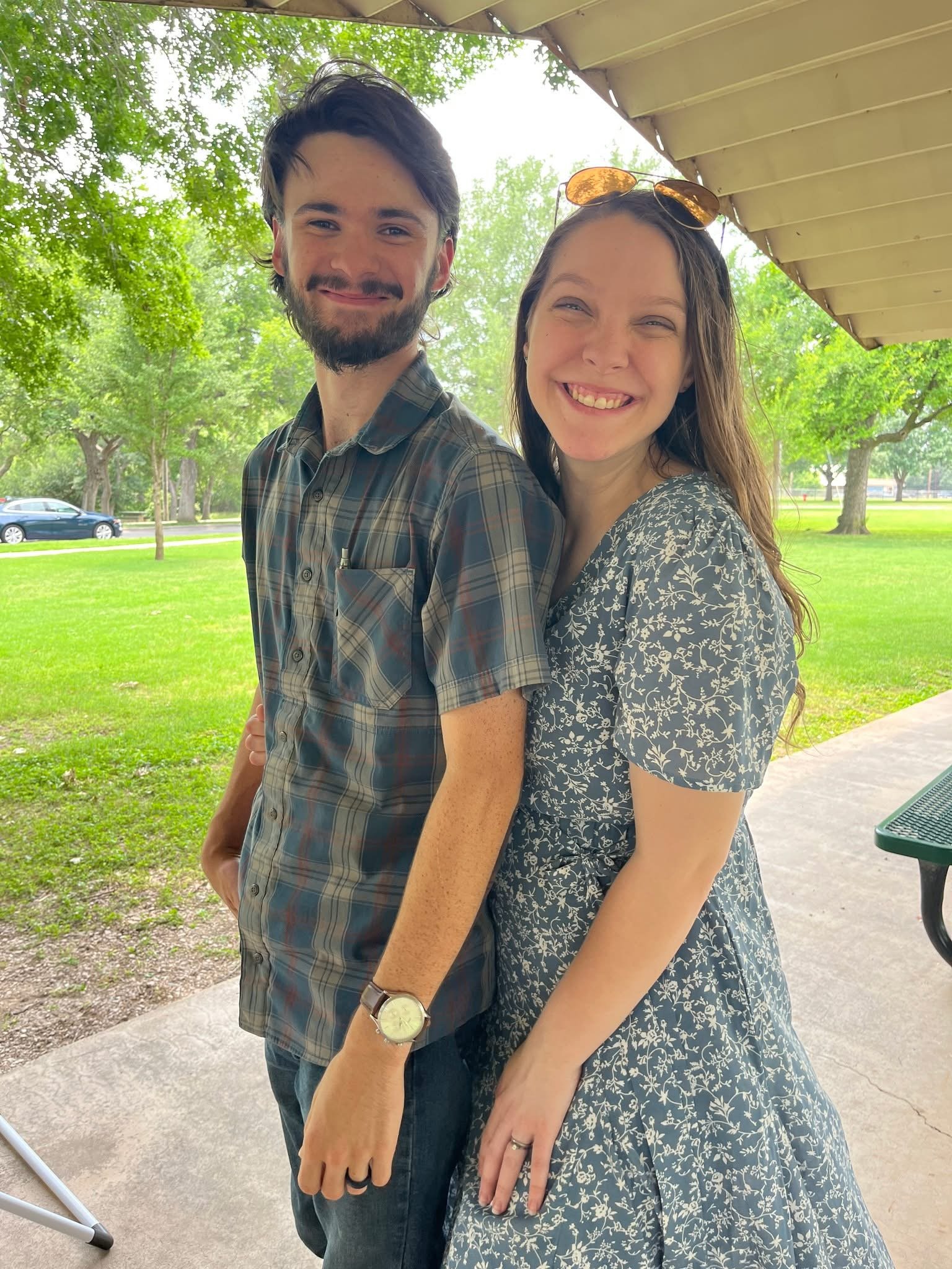 A smiling young man and woman standing outdoors under a roof, with a park and trees in the background.