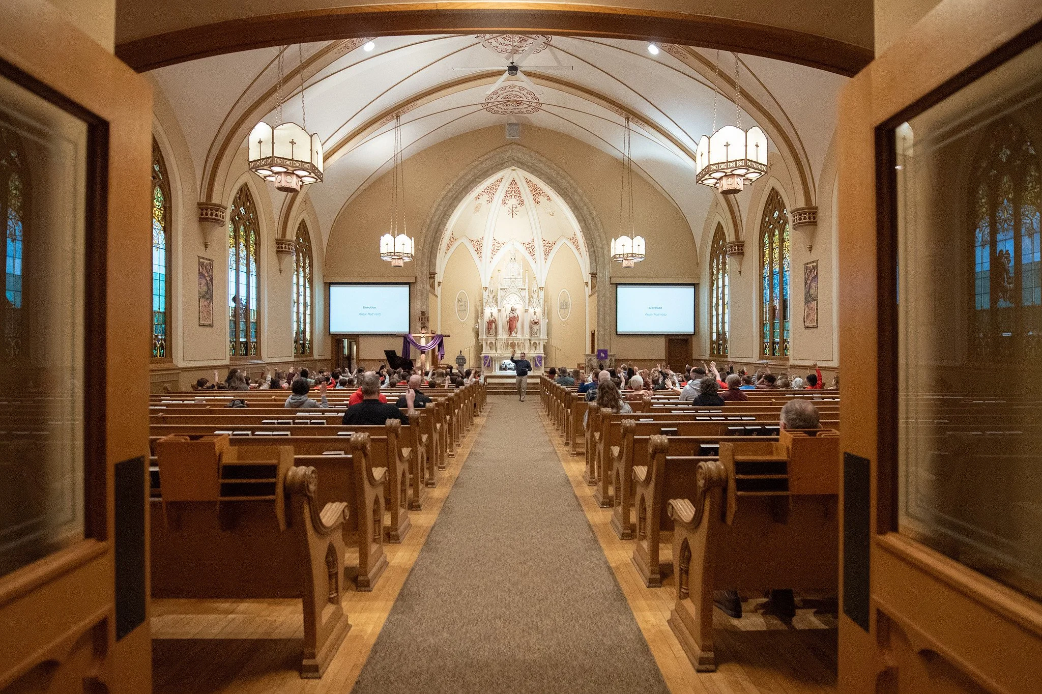 Looking into the interior of a church with wooden pews, stained glass windows, and an altar with religious statues, viewed through a partially opened door.