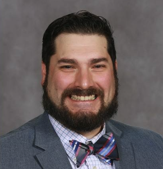 A man with dark hair and a beard, smiling, wearing a blue suit, checkered shirt, and colorful bow tie, against a gray background.