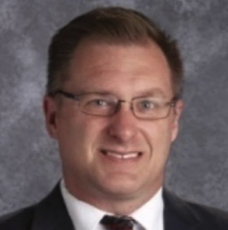 A man wearing glasses and a suit smiling in front of a gray background.