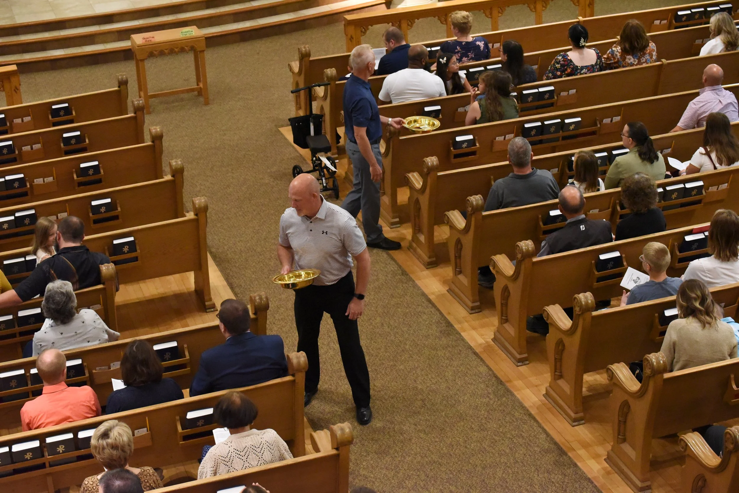People seated in wooden pews inside a church, some holding programs, while two men carry gold trays through the aisle.