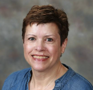 A woman with short brown hair, smiling, wearing a blue top, against a blurred gray background.
