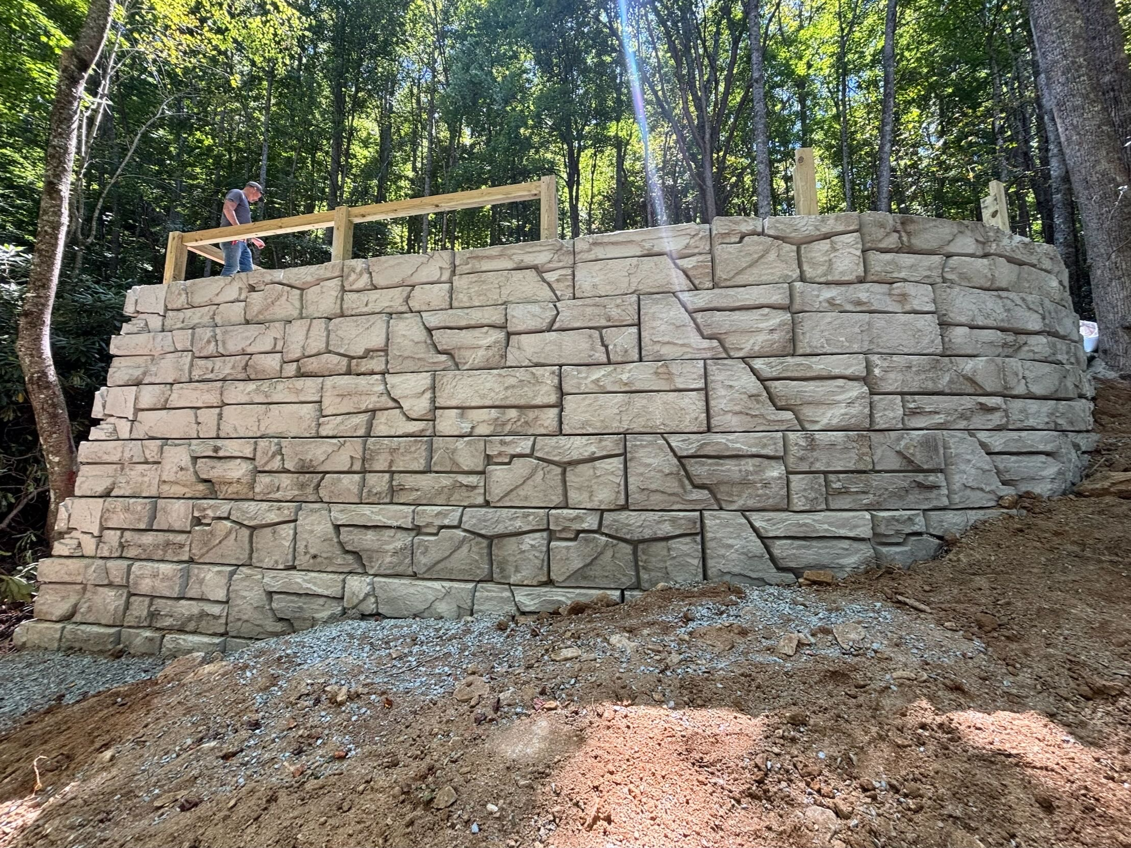 A stone retaining wall under construction in a wooded area with two workers on top of the wall working on the wooden railing.