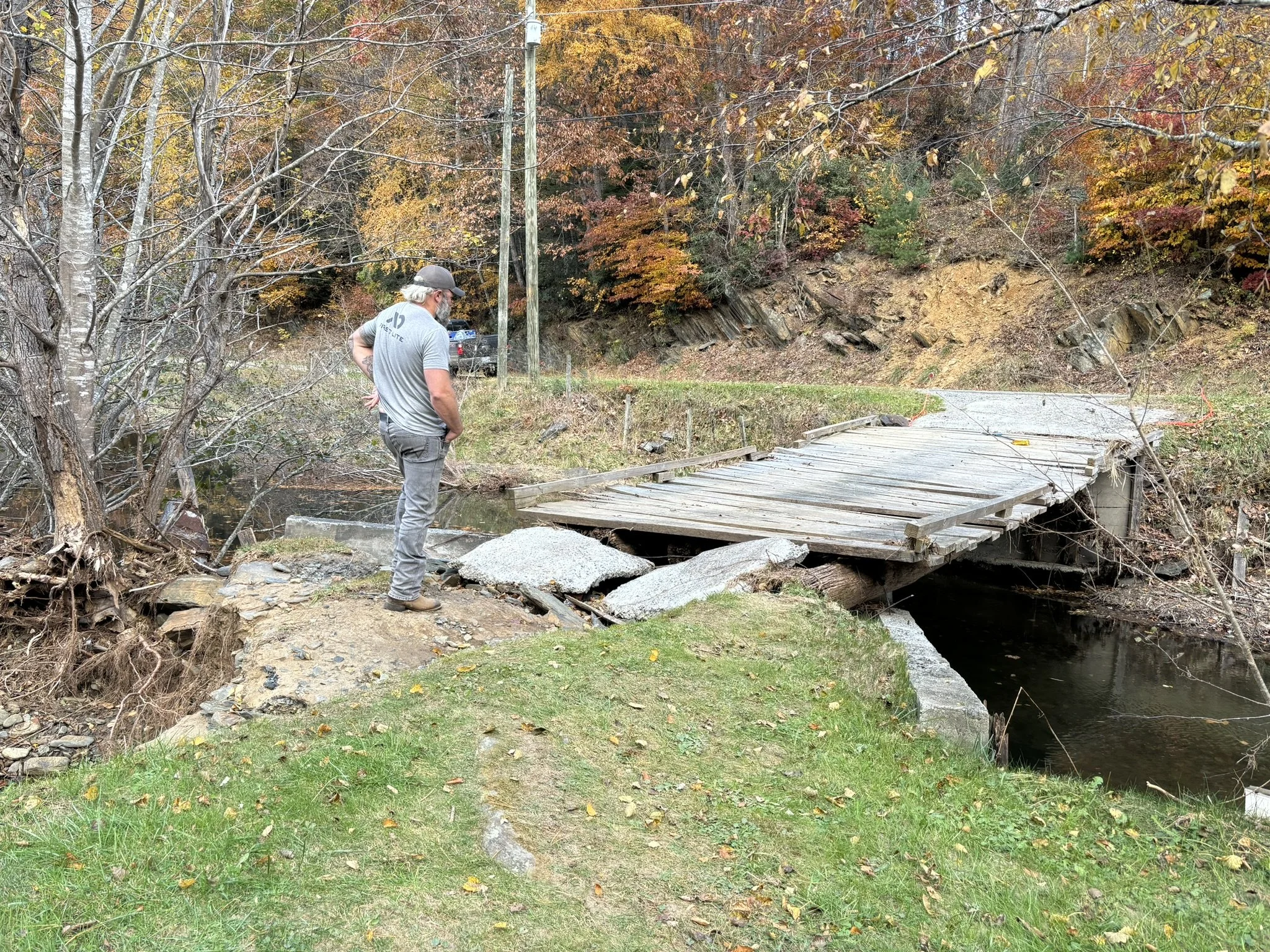 A man standing on a grassy area near a damaged wooden bridge over a small creek, with autumn trees and electrical poles in the background.