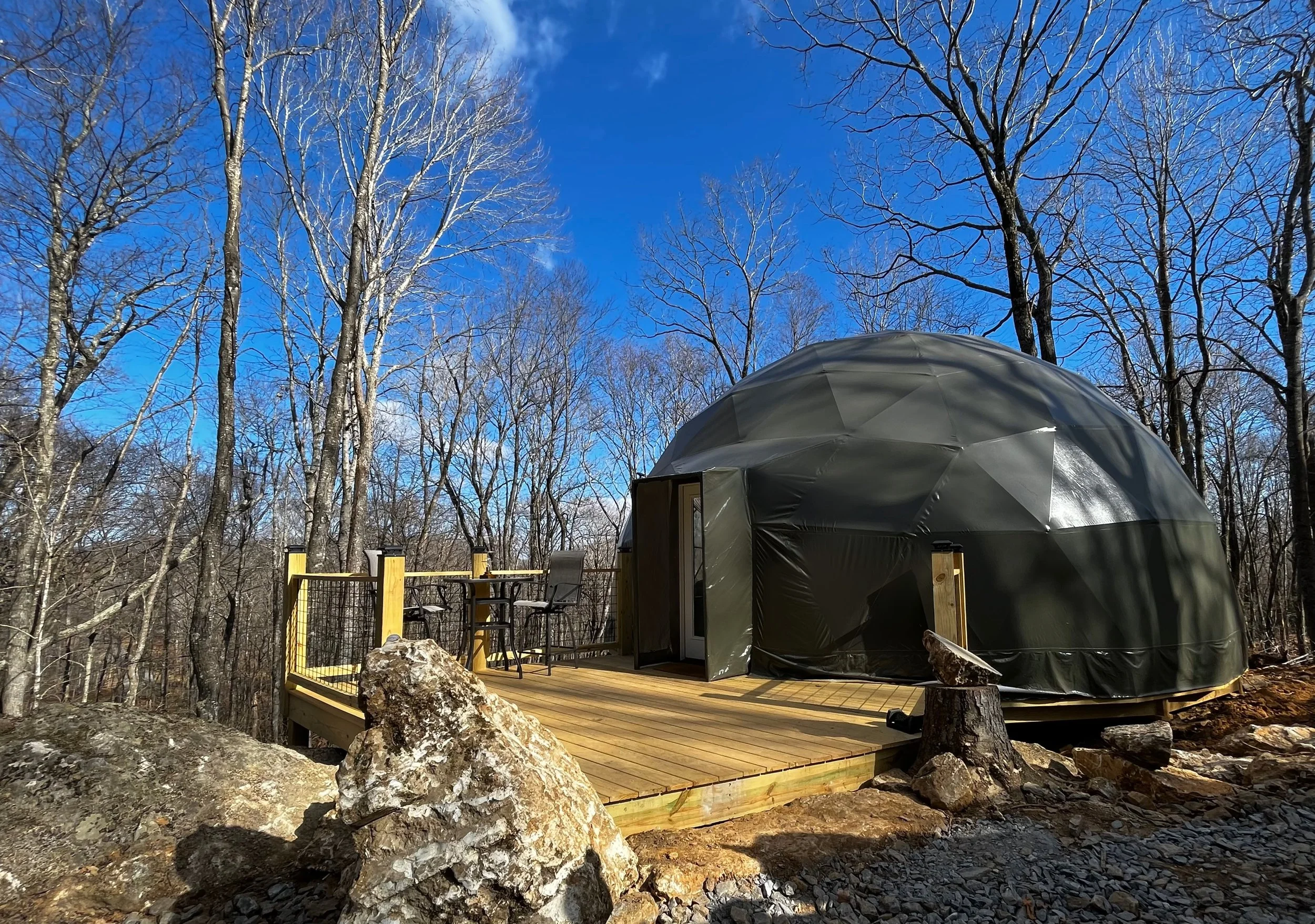 A geodesic dome cabin on a wooden deck in a forest with leafless trees and a blue sky.