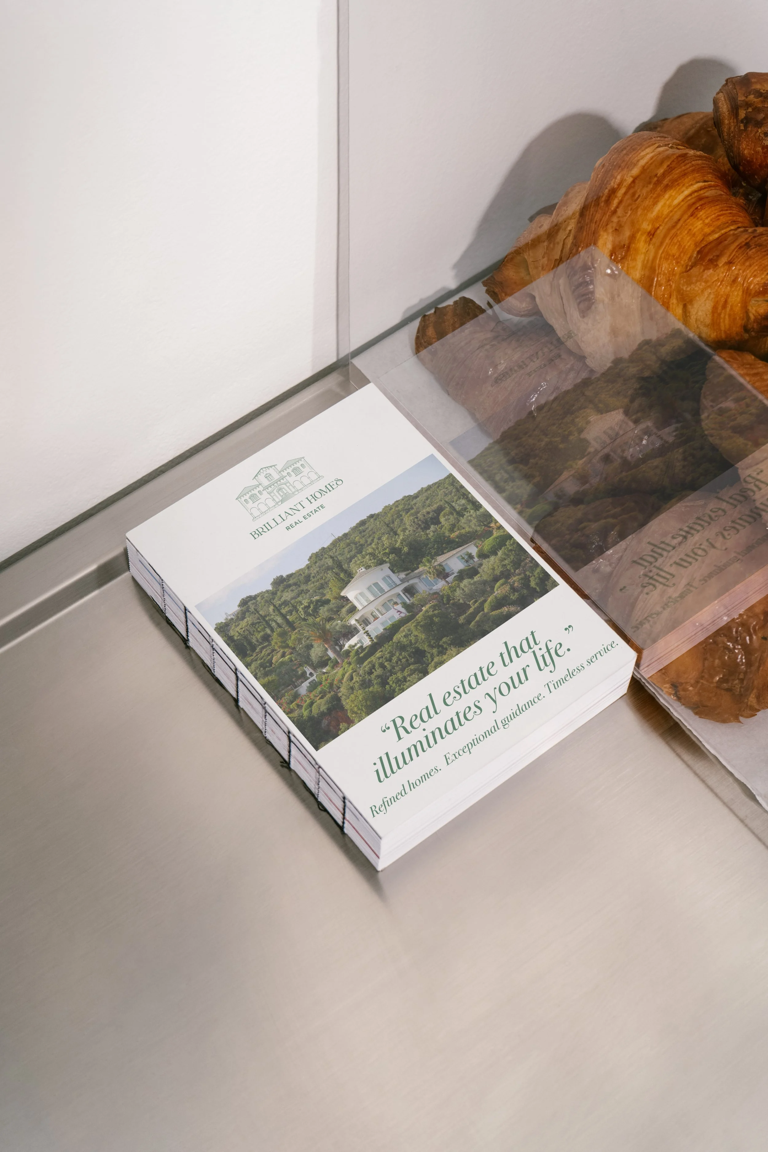 Folleto de bienes raíces en una mesa, con imagen de una casa en un entorno natural y texto que dice 'Real estate that illuminates your life' y 'Refined homes. Exceptional guidance. Timeless service'.