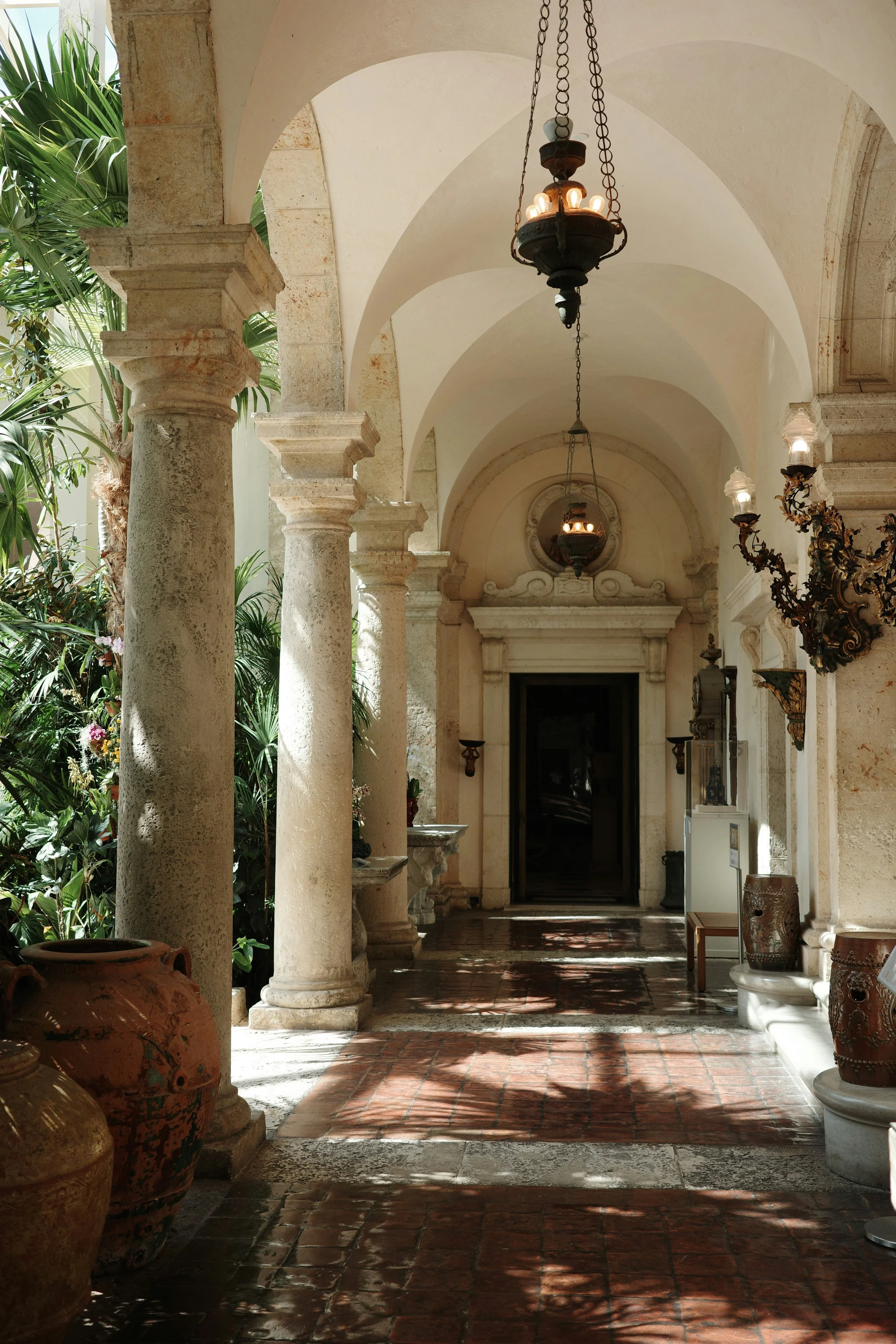 Pasillo cubierto con columnas de piedra, lámparas colgantes y macetas de cerámica, rodeado de plantas verdes y decoraciones antiguas.