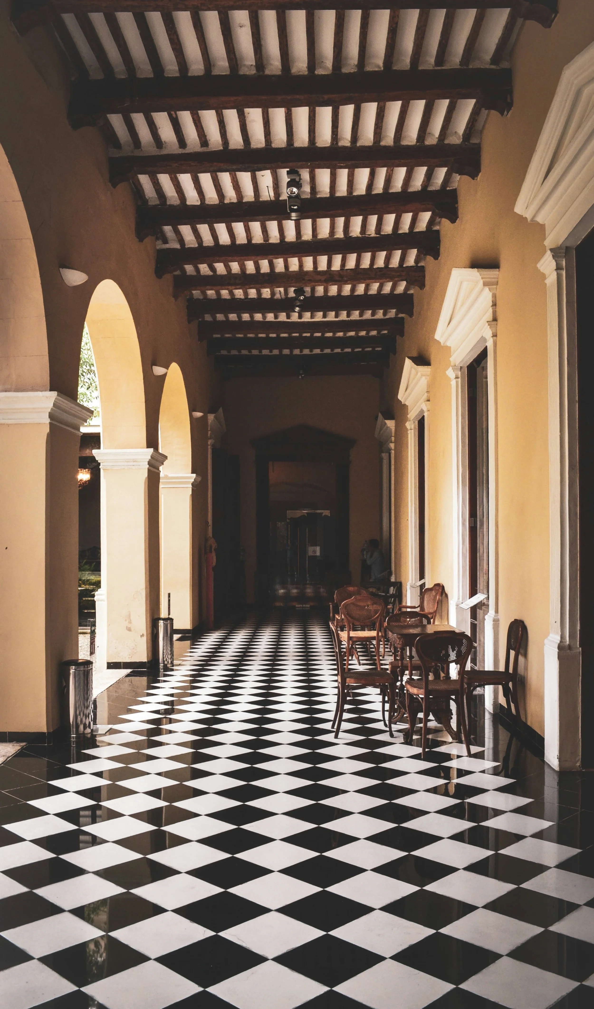 Pasillo interior con piso de azulejos en patrón de cuadros blancos y negros, paredes de color beige, arcos y ventanas con molduras blancas, muebles de madera en el lado derecho, techo con vigas de madera expuestas y lámparas en el techo.