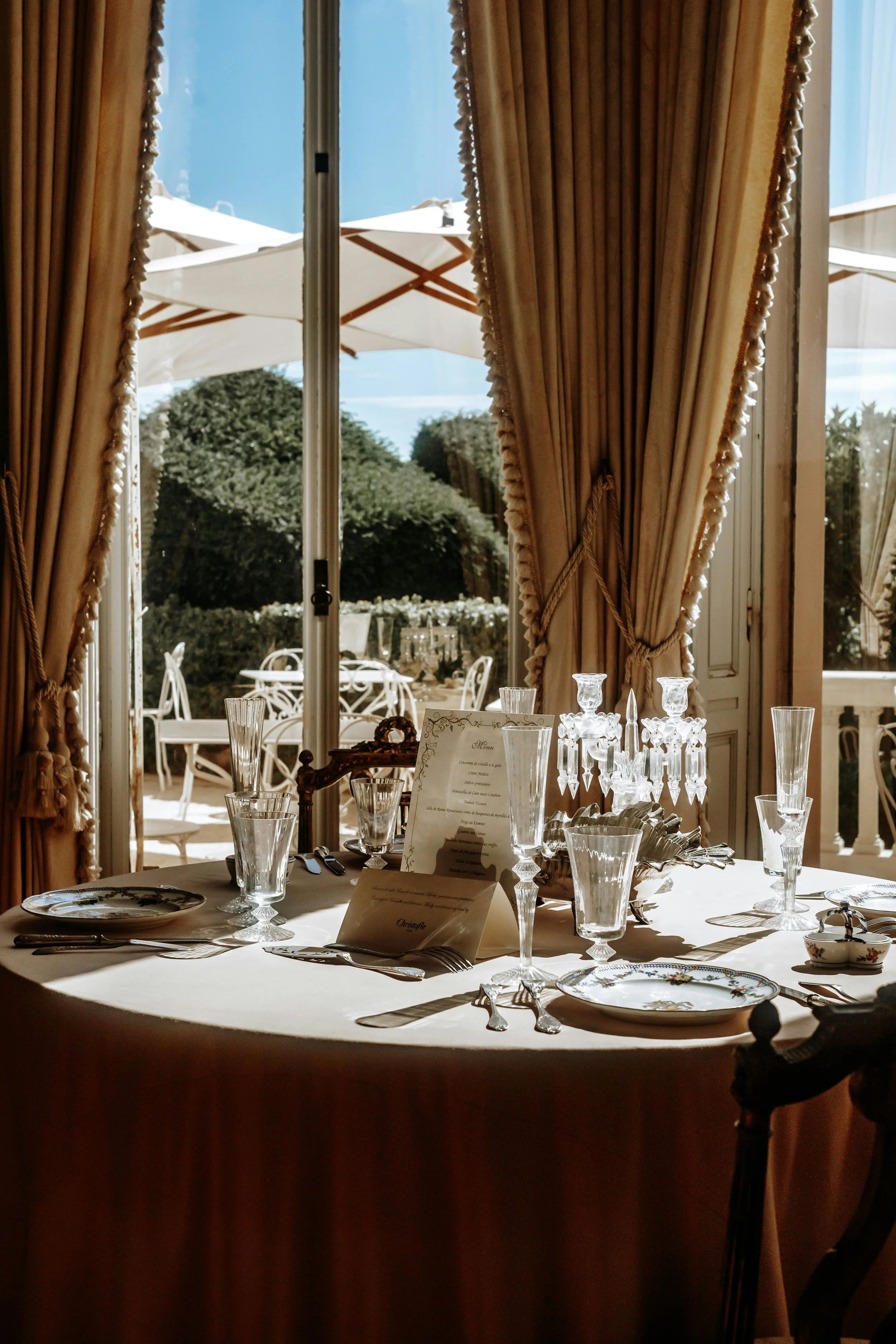 Mesa elegant decorada para una comida o evento, con platos, copas y cubiertos, en un ambiente con cortinas doradas y vista exterior con sombrillas y mobiliario blanco.