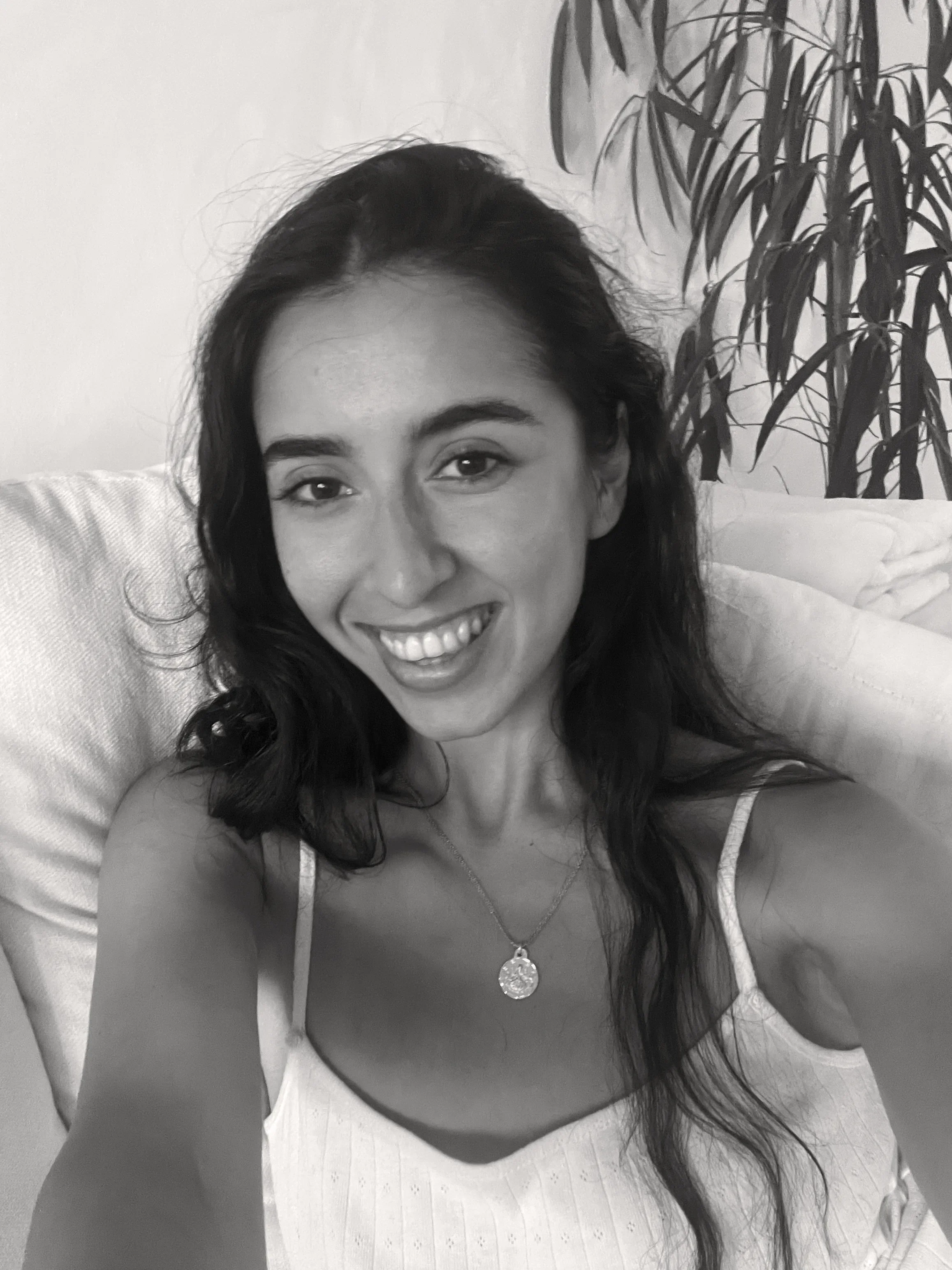 Mujer sonriendo, con cabello oscuro y vestida con blusa blanca, en un entorno doméstico con planta en el fondo.