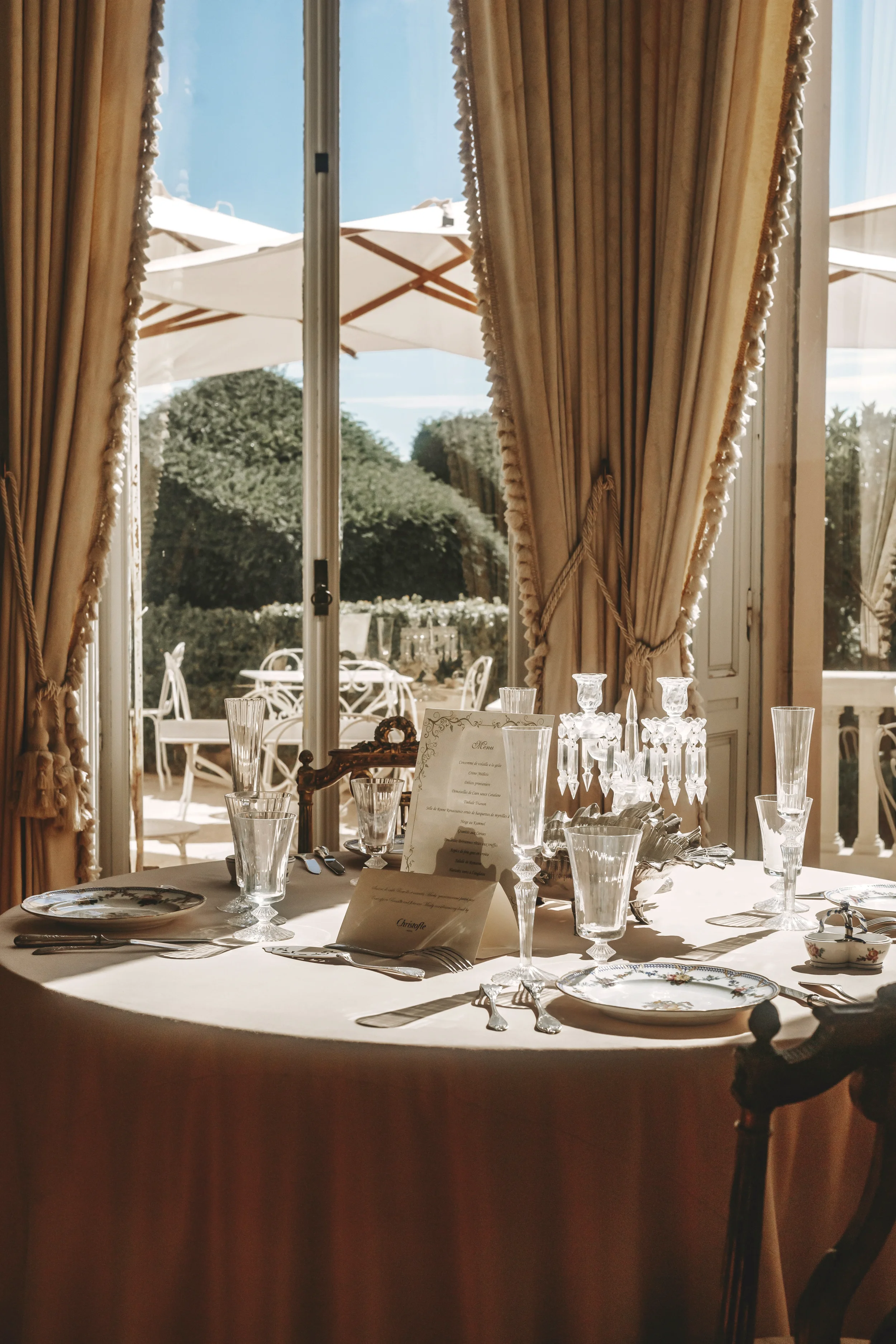 Mesa decorada con vajilla, cristalería, mantelería y un candelabro, en una habitación con cortinas doradas y vista al exterior con sillas y mesas en un jardín bajo paraguas blancos.