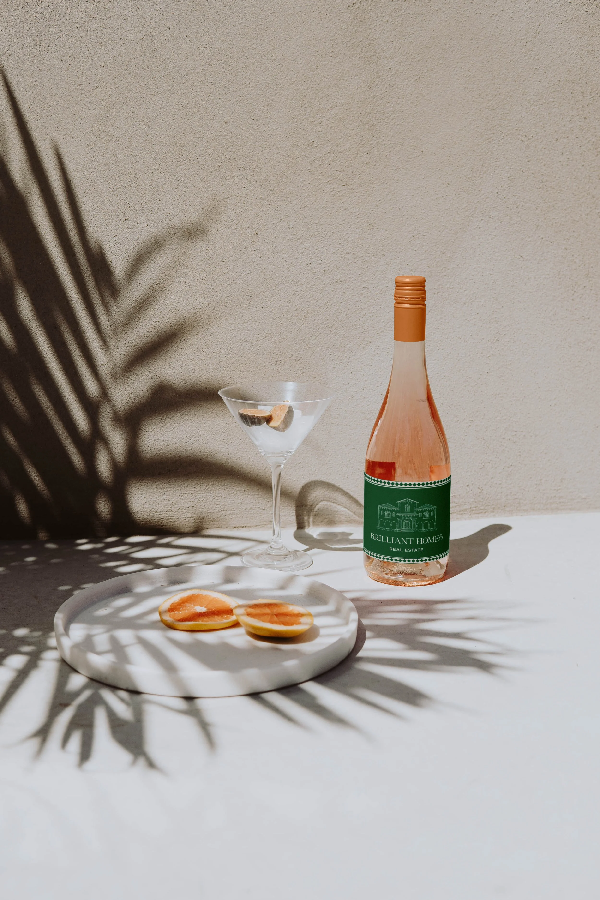 Botella de vino rosado, copa de cóctel con rodajas de fruta, y rebanadas de naranja sobre una bandeja, con sombras de plantas en un fondo claro.