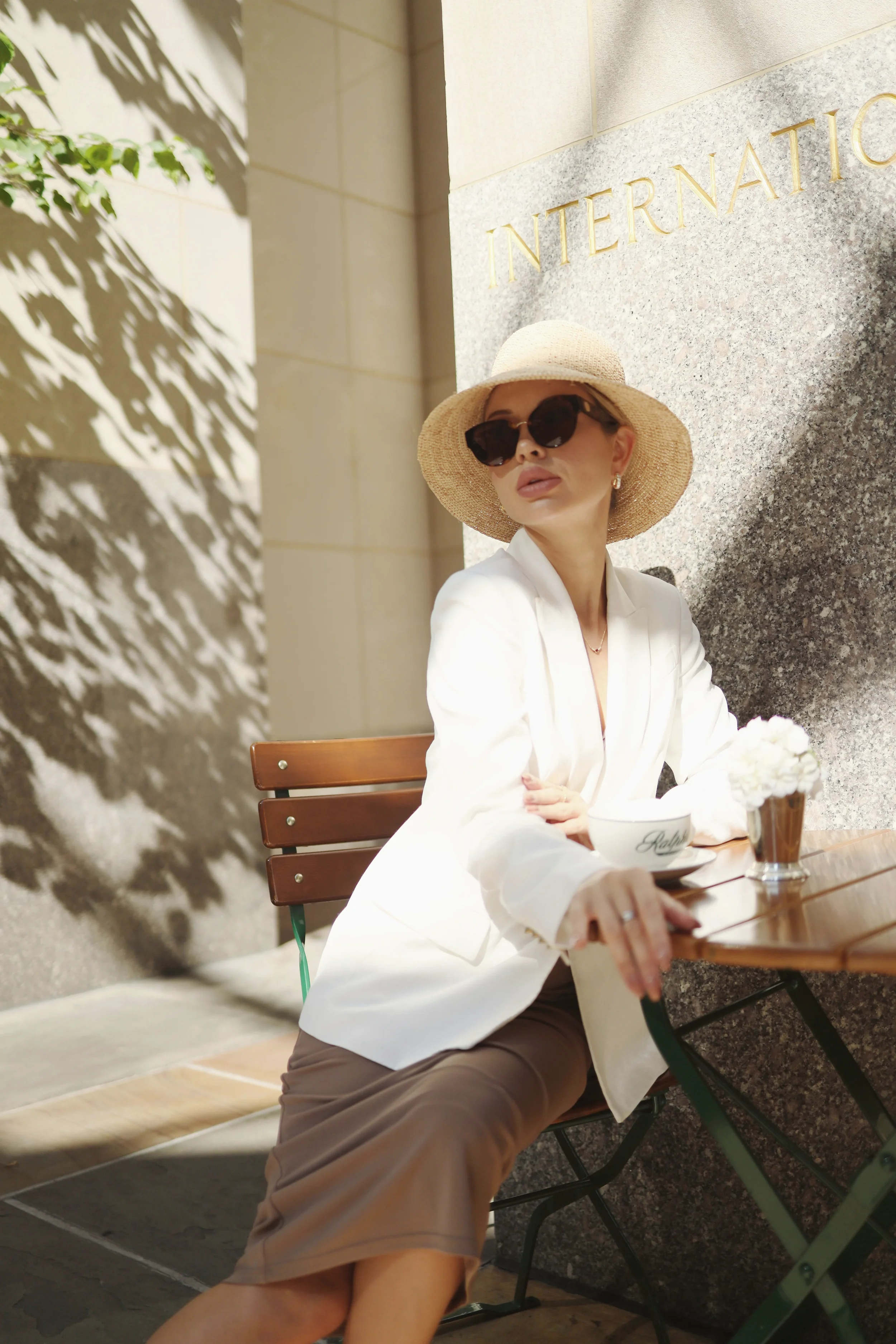 Mujer elegante con sombrero de paja y gafas oscuras, sentada en una mesa de madera con una taza, un florero con flores blancas y un fondo de pared de piedra con la palabra 'INTERNATIONAL' en letras doradas.