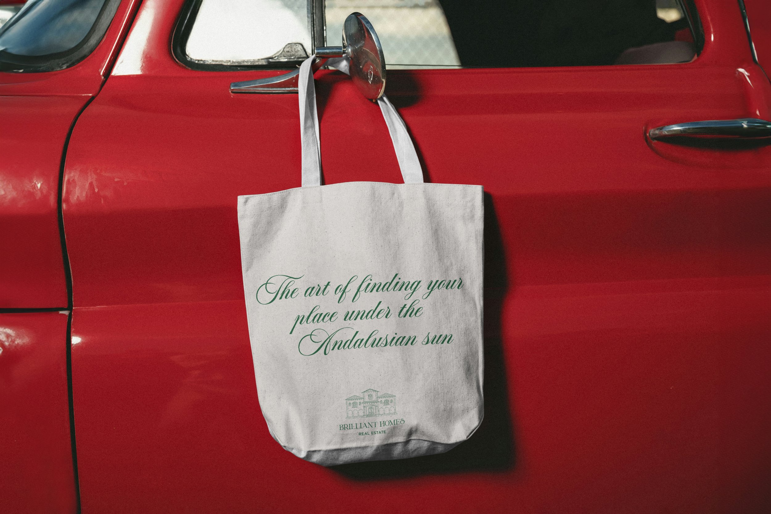 Bolsa de tela colgada en el espejo retrovisor de un coche rojo, con la frase "El arte de encontrar tu lugar bajo el sol andaluz".