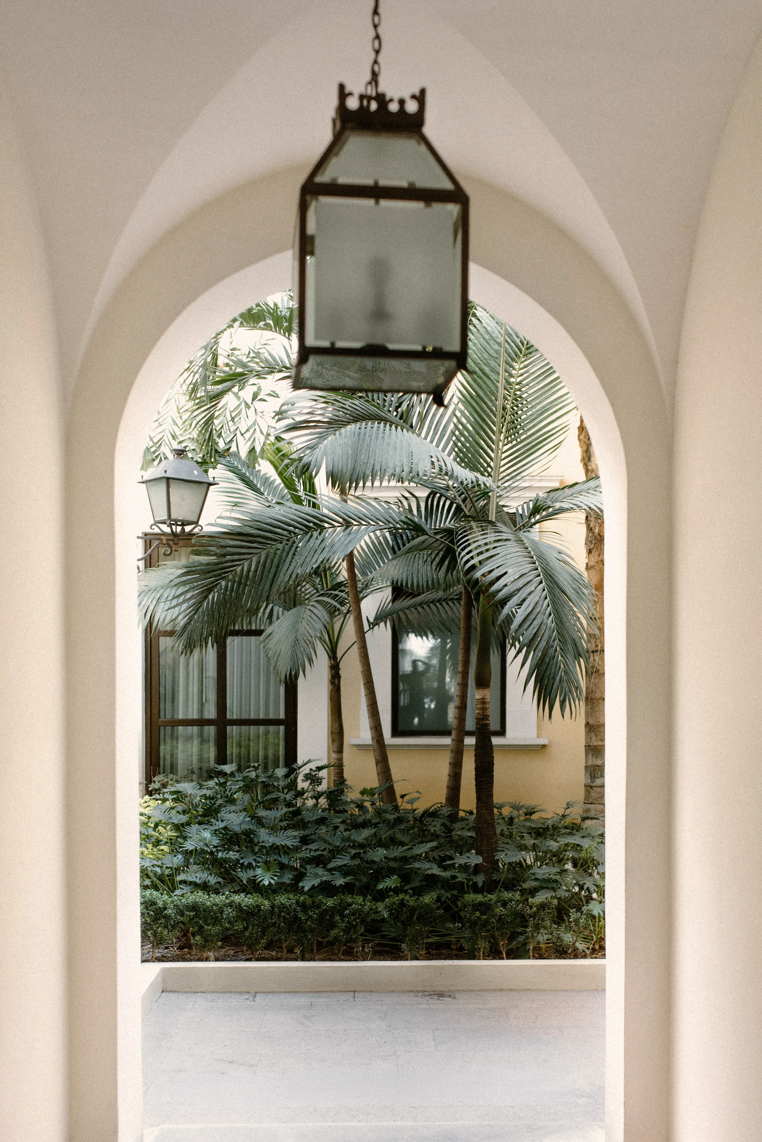 Vista de un patio con plantas tropicales, desde un arco con lámpara colgante en primer plano