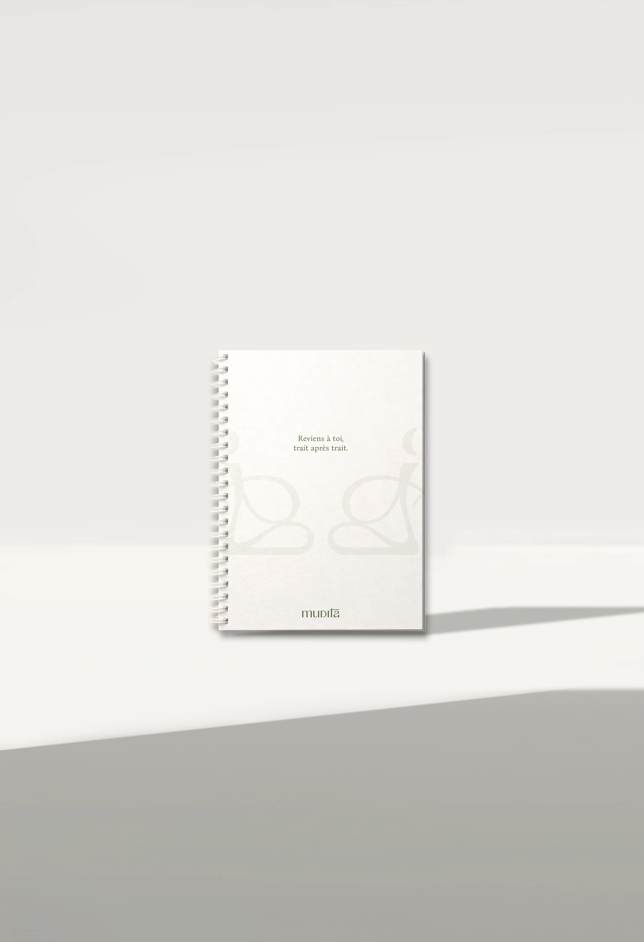 Cuaderno de tapa blanca con impresión sutil y espiral en el lado izquierdo, mostrando en la portada las palabras 'Reviens à toi, trait après trait' y en la parte inferior la marca 'mudita', colocado sobre una superficie blanca con sombra suave.
