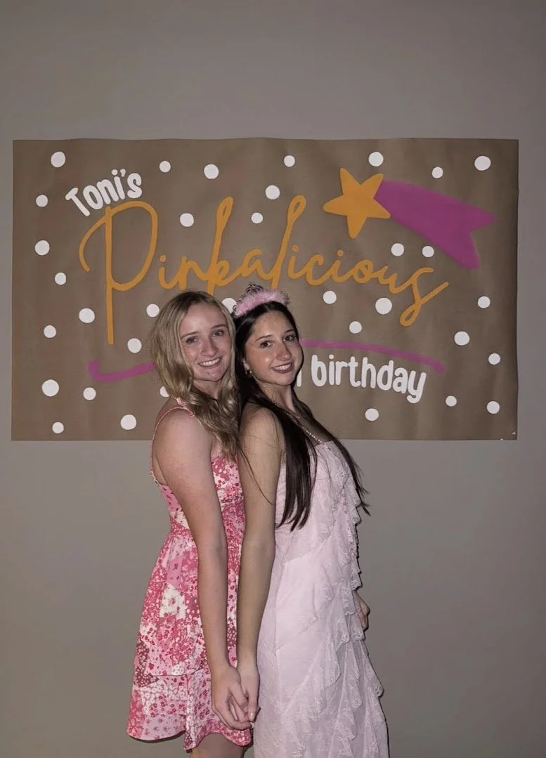 Two young women at a birthday party standing in front of a banner that says "Toni's Pinkalicious birthday," one wearing a pink dress and the other wearing a light pink dress with a pink headband.
