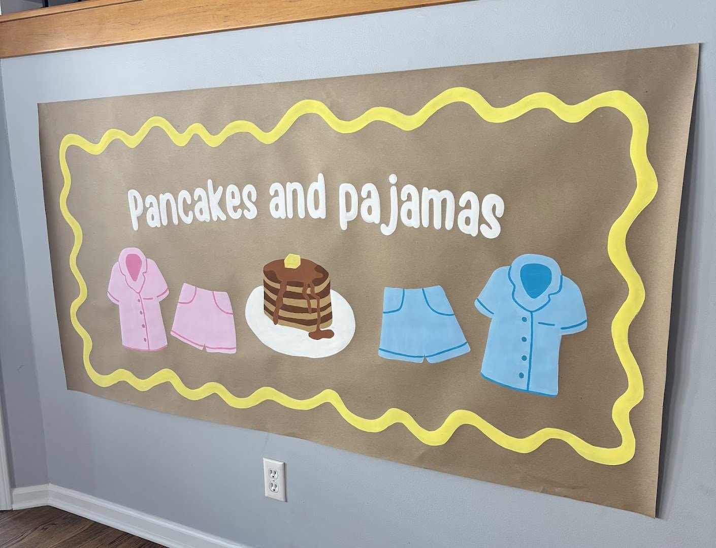 Decorative banner with the phrase 'Pancakes and pajamas' and illustrations of pink and blue pajamas, a stack of pancakes with syrup, and a yellow wavy border.