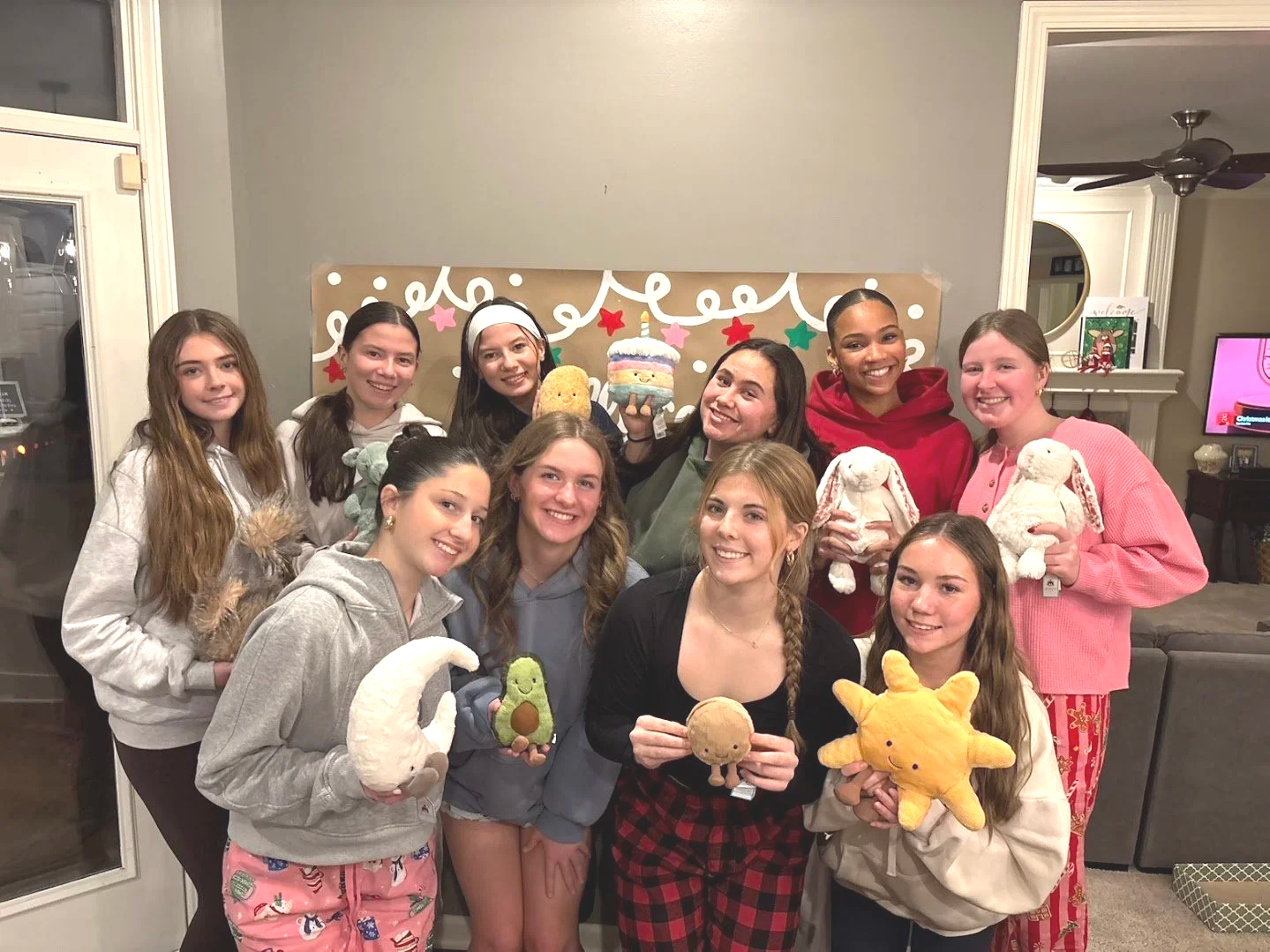 A group of twelve young women gathered indoors, smiling and holding plush toys, pajamas, and festive decorations.
