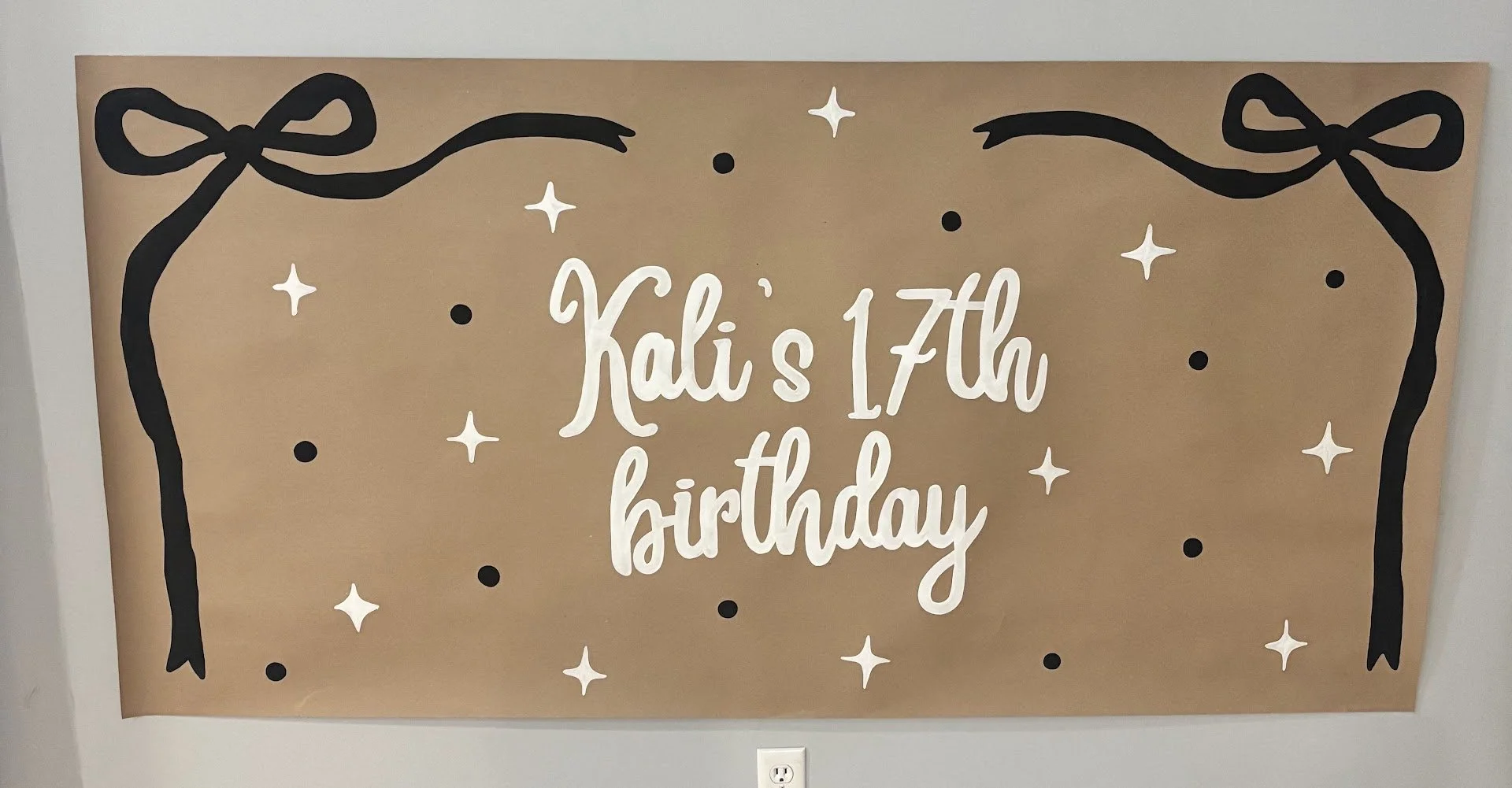 Decorative birthday sign with black bows, white stars, and black dots, saying 'Kali's 17th birthday' in white cursive letters on brown paper background.