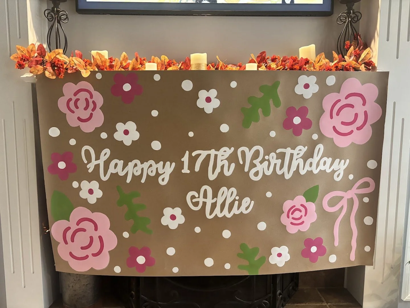 Decorative birthday sign with pink and white flowers, green leaves, and a pink ribbon bow. The sign reads 'Happy 17th Birthday Allie' in white cursive letters. The top features orange, red, and pink artificial flowers and candles.
