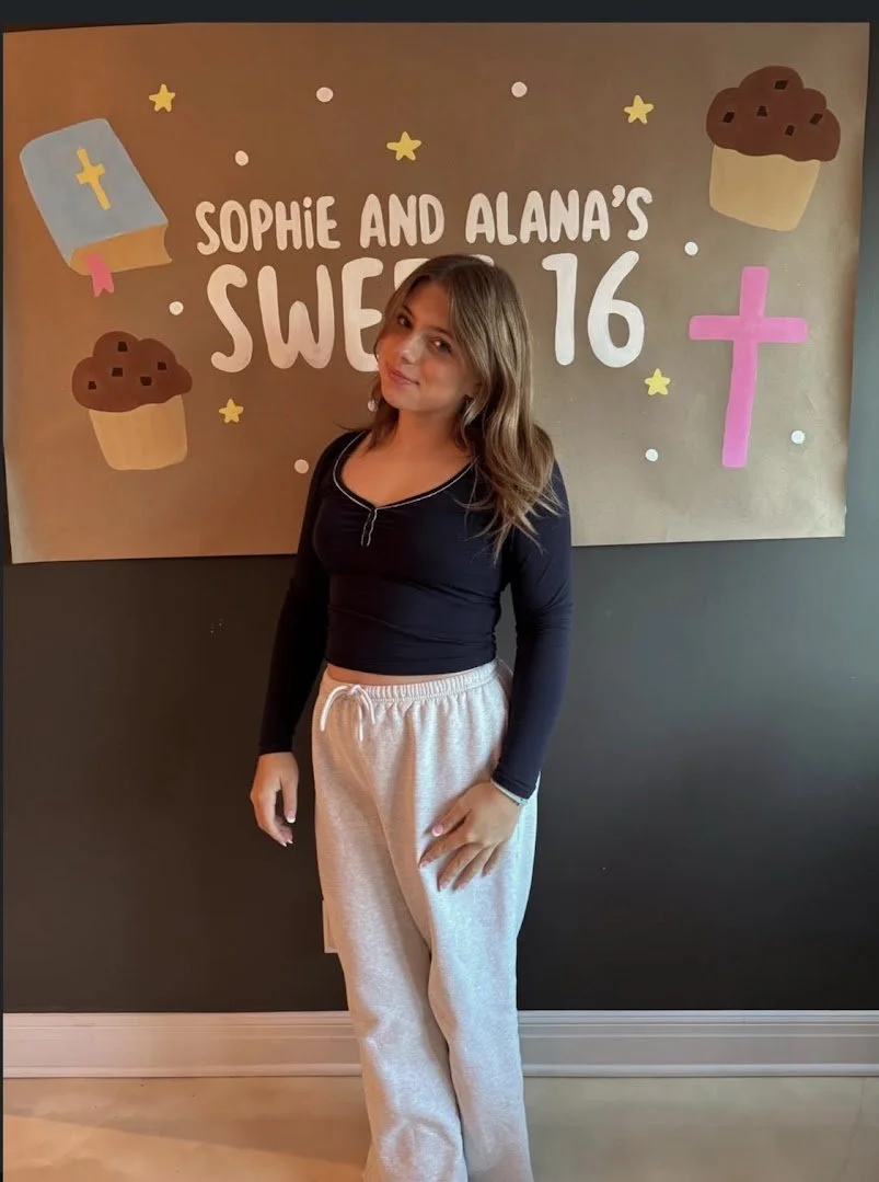 Young girl at a sweet 16 birthday party, standing in front of a decorated backdrop with cupcakes, a cross, and stars, smiling slightly and wearing a black long-sleeve top and gray sweatpants.