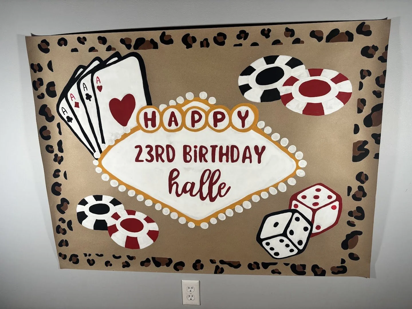 A handmade birthday poster featuring a casino theme with playing cards, poker chips, and dice. The message reads, 'Happy 23rd Birthday Halle,' with the number 23 circled and decorated, set against a background of leopard print border.