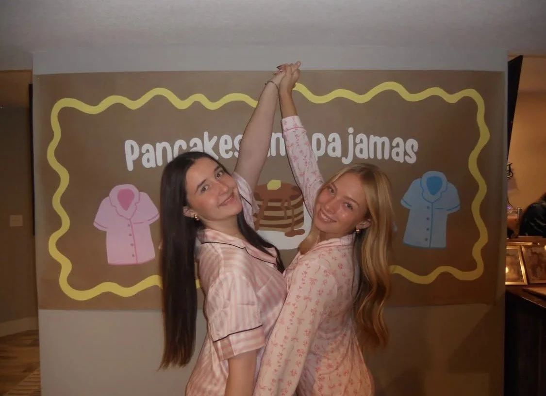Two women in pink pajamas celebrating in front of a backdrop with the words "Pancakes or Pajamas" and icons of pajamas and pancakes, holding hands above their heads and smiling.