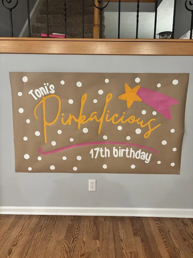 Decorative sign for a 17th birthday party with the words "Toni's Pinkalicious" signed in orange, pink, and white, surrounded by white polka dots, a pink shooting star, and a pink trail on a brown paper background.