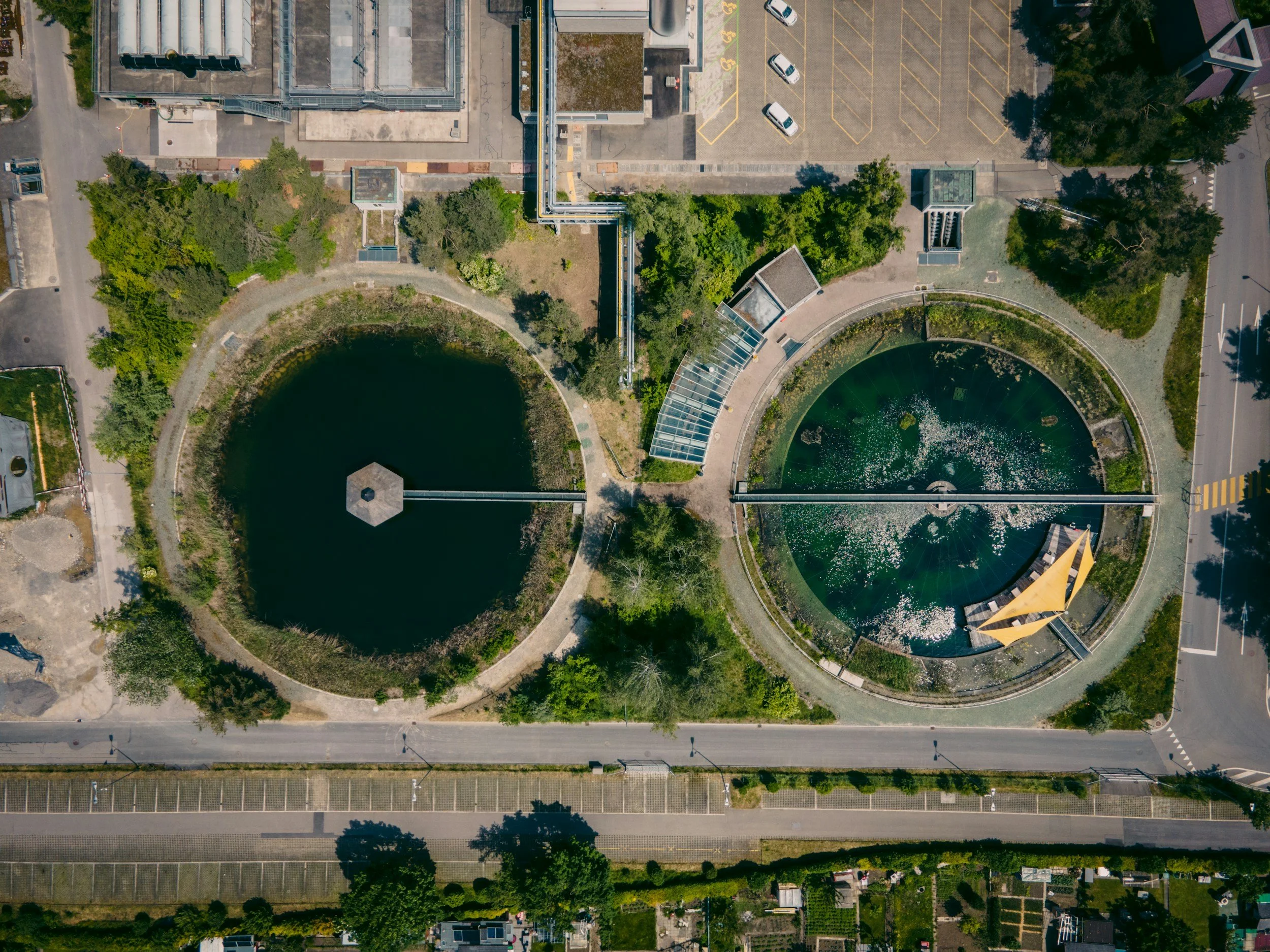 Aerial view of two circular water features in an urban park, with greenery, pathways, and surrounding buildings, one with a hinged yellow sculpture or structure.