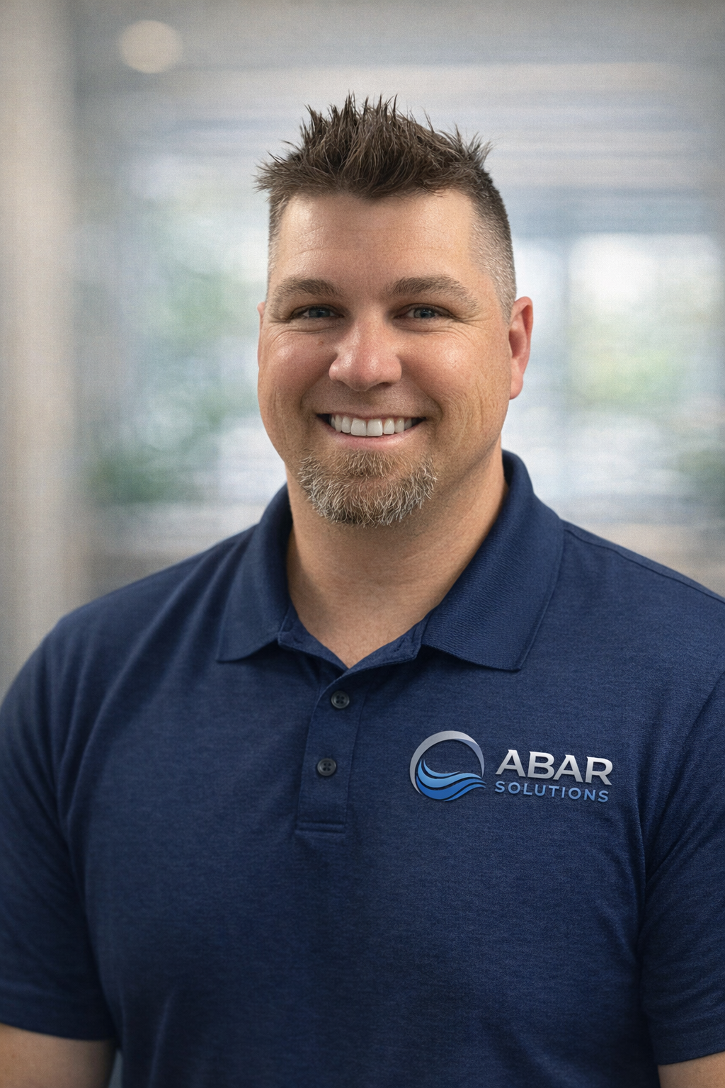 Smiling man with short spiky hair, goatee, wearing a navy blue polo shirt with 'ABAR Solutions' logo, sitting indoors with blurred window background.