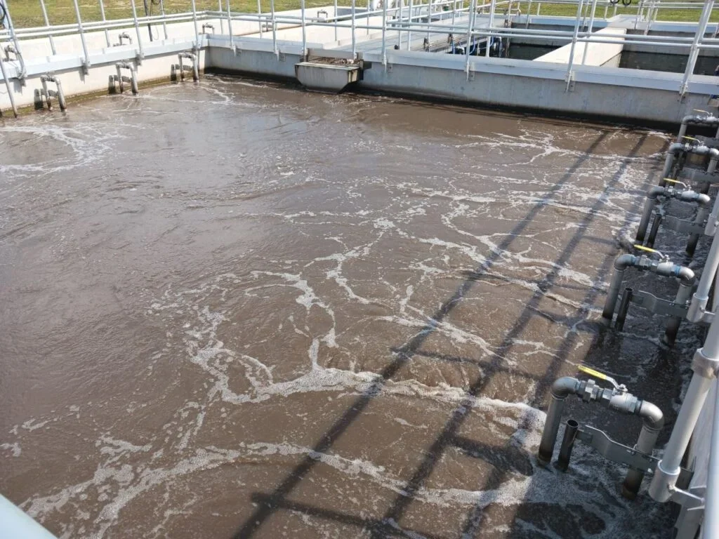Water treatment plant's aeration basin with foamy water and pipes.