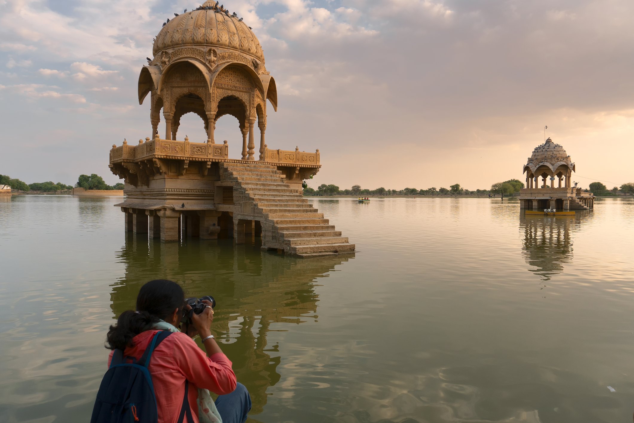 A person with a backpack taking a photo of two historic stone pavilions on a lake, with one in the foreground and one in the background, under a partly cloudy sky.